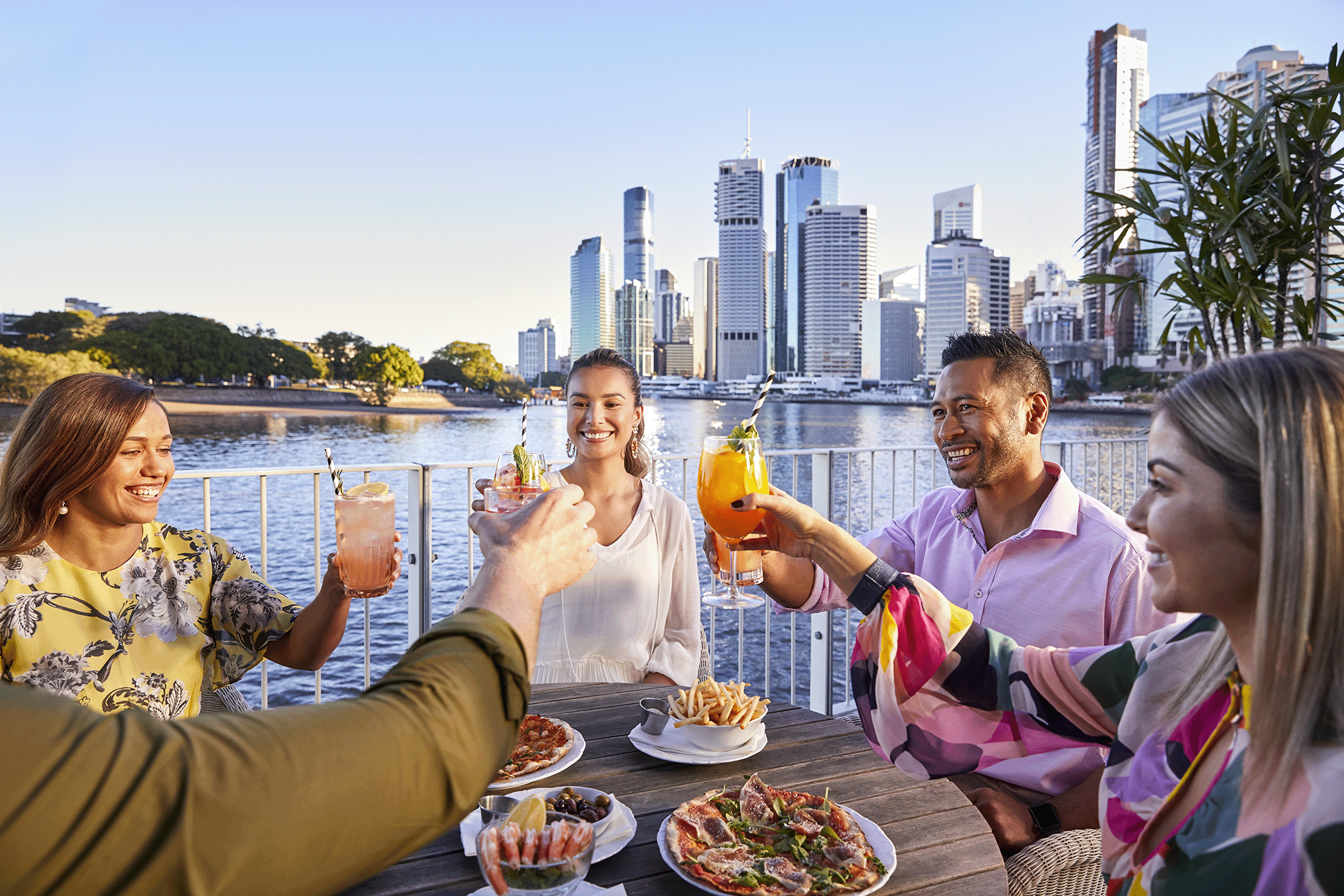 A group of friends alfresco dining and cheering over cocktails at riverside bar Brisbane City